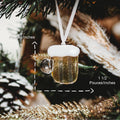 A miniature beer mug Christmas ornament hanging from a white ribbon, with overlaid dimension arrows and text showing 2 pouces/ingles tall and 1 1/2 pouces/ingles wide, against a blurred Christmas tree background.