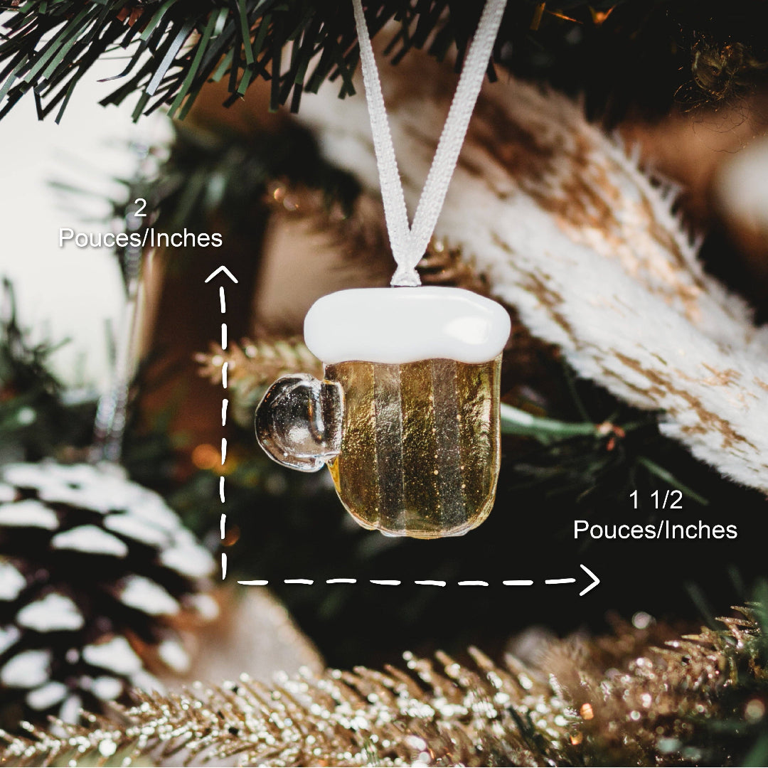 A miniature beer mug Christmas ornament hanging from a white ribbon, with overlaid dimension arrows and text showing 2 pouces/ingles tall and 1 1/2 pouces/ingles wide, against a blurred Christmas tree background.