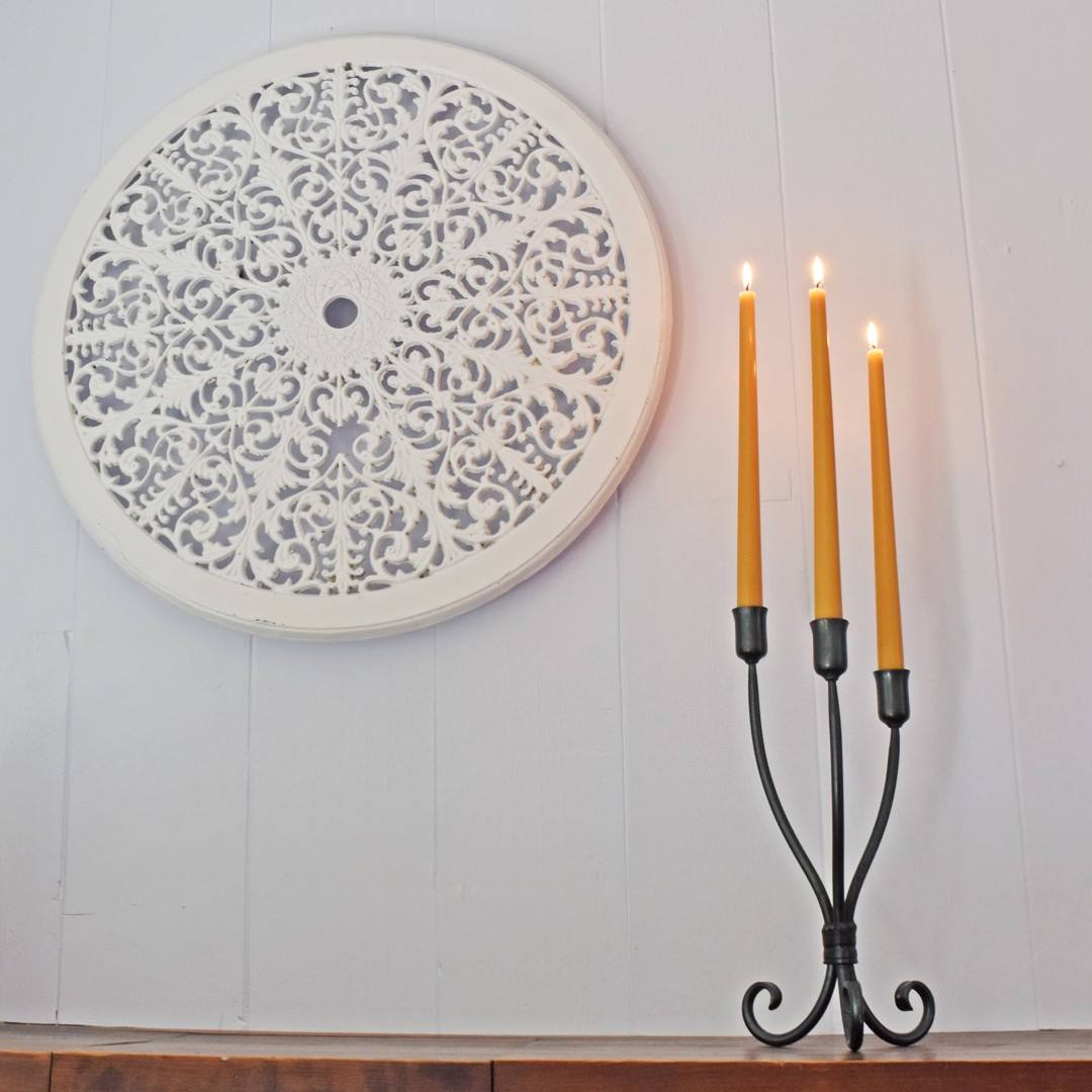 A white, round wall hanging with a detailed, intricate pattern hangs above a black wrought-iron candlestick holding three lit yellow candles.