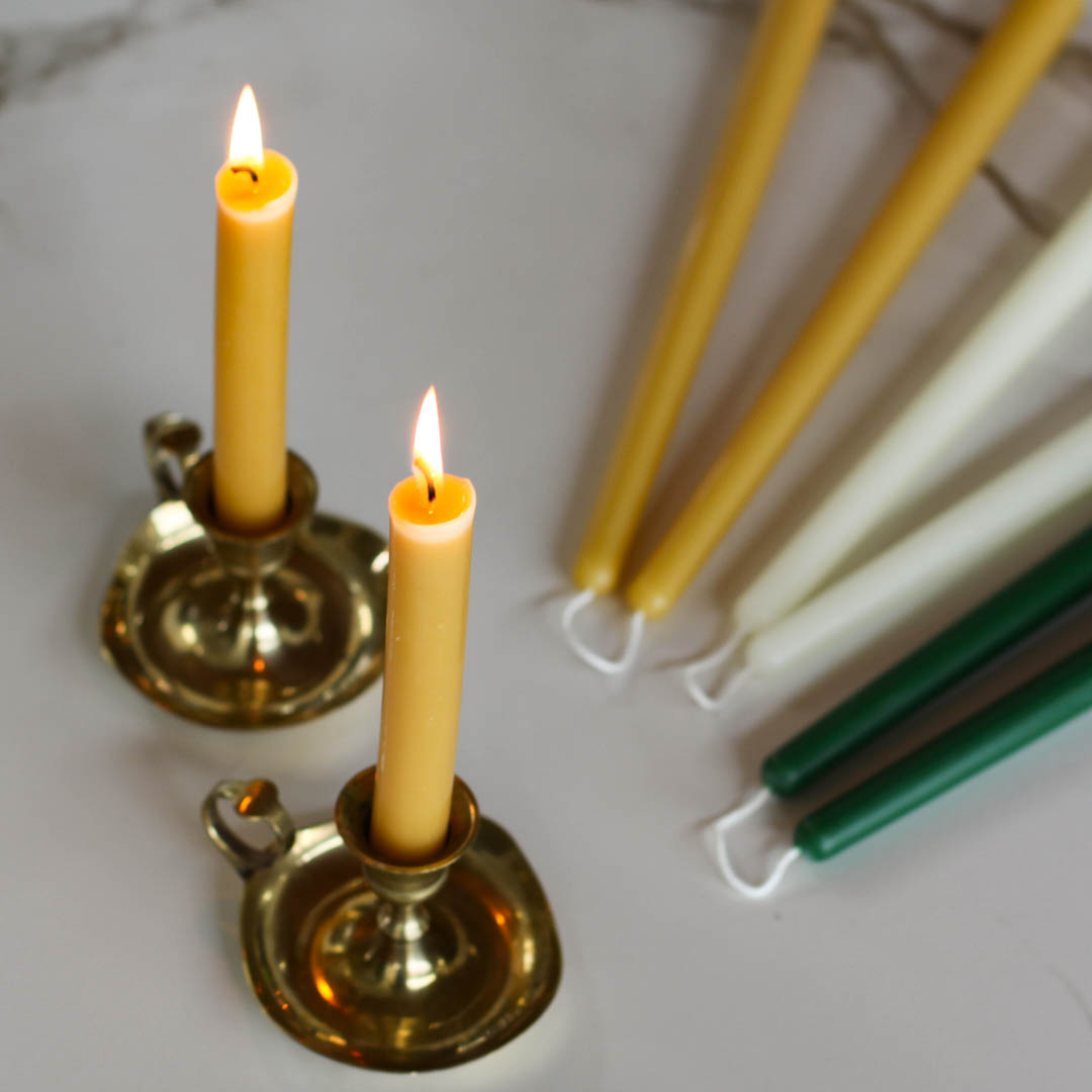 Two lit beeswax candles in brass candle holders, surrounded by several unlit beeswax candles in various sizes and colors, including yellow, cream, and green.