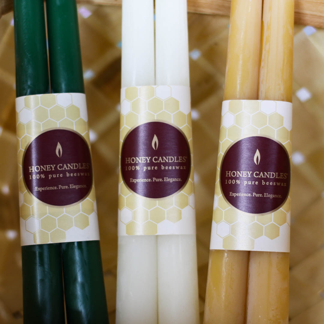 Three pairs of beeswax candles in different colors, including green, cream, and yellow. The candles are bundled together with labels that say "HONEY CANDLES 100% Pure Beeswax" and a honeycomb pattern.