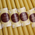 Multiple rows of yellow beeswax candles, each with a label that says "HONEY CANDLES 100% Pure Beeswax" and a honeycomb pattern. The candles are arranged tightly together in a grid-like pattern.