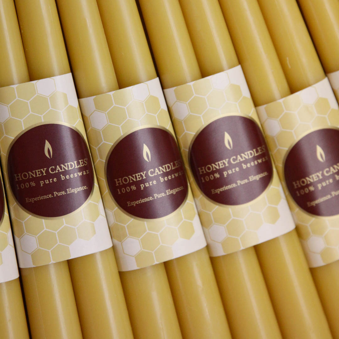 Multiple rows of yellow beeswax candles, each with a label that says "HONEY CANDLES 100% Pure Beeswax" and a honeycomb pattern. The candles are arranged tightly together in a grid-like pattern.
