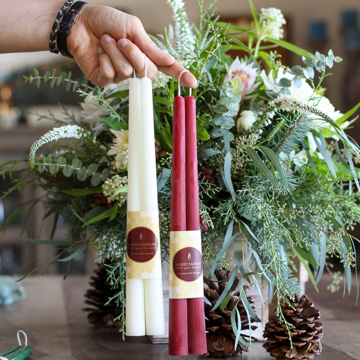 A person's hand holding two pairs of beeswax candles, one pair white and one pair maroon. The candles are standing upright and have labels that say 