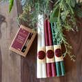 A collection of beeswax candles on a wooden table. There is a box of beeswax tealights, as well as several longer beeswax candles in cream, maroon, and green. The candles are surrounded by greenery and flowers.