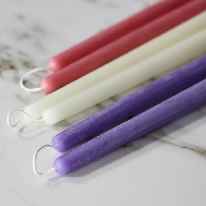 A close-up of several beeswax candles in various colors, including pink, cream, and purple. The candles are arranged side by side on a white marble surface.