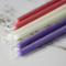 A close-up of several beeswax candles in various colors, including pink, cream, and purple. The candles are arranged side by side on a white marble surface.