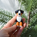 A hand holds a small dog-shaped Christmas ornament against a background of green pine branches. The ornament is shaped like a Bernese mountain dog with a black, white, and orange color scheme and hangs from a white ribbon.