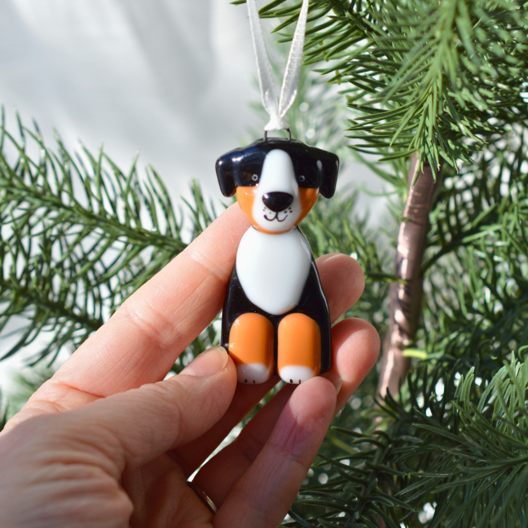 A hand holds a small dog-shaped Christmas ornament against a background of green pine branches. The ornament is shaped like a Bernese mountain dog with a black, white, and orange color scheme and hangs from a white ribbon.