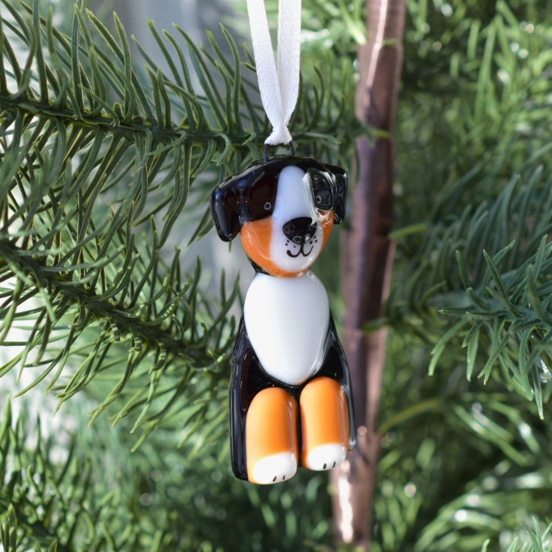 A small dog-shaped Christmas ornament hanging from a white ribbon on a green evergreen tree branch. The dog has black, white, and orange coloring resembling a Bernese mountain dog.
