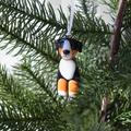A small dog-shaped Christmas ornament hanging from a white ribbon among green pine tree branches. The ornament is black, white, and orange, styled like a Bernese mountain dog.