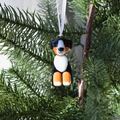 A small dog-shaped Christmas ornament hanging from a white ribbon on a green evergreen tree branch. The dog has black, white, and orange coloring resembling a Bernese mountain dog.