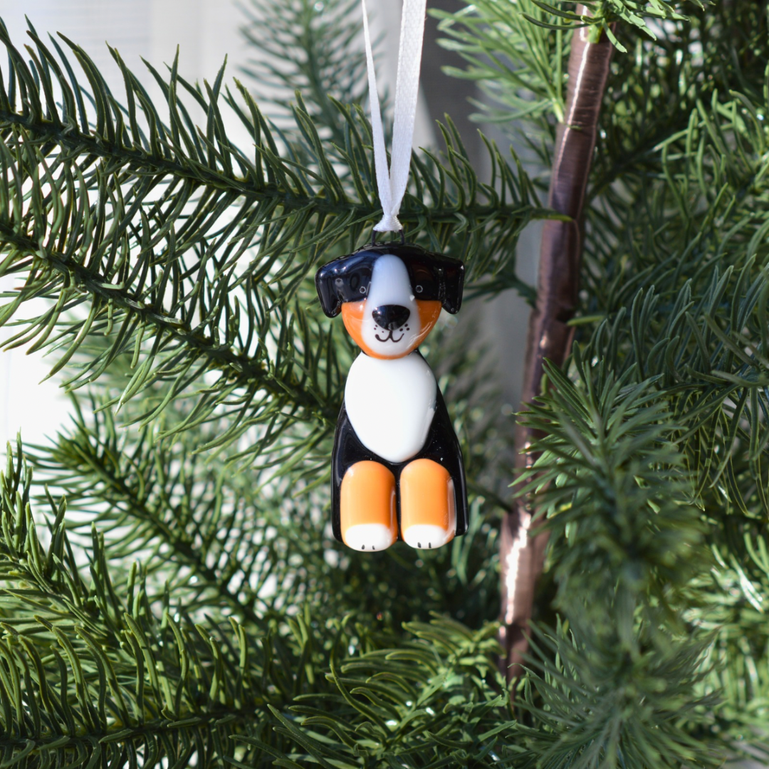 A small dog-shaped Christmas ornament hanging from a white ribbon on a green evergreen tree branch. The dog has black, white, and orange coloring resembling a Bernese mountain dog.