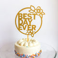 Gold 'Best Day Ever' cake topper on a white cake with colorful sprinkles against a white background