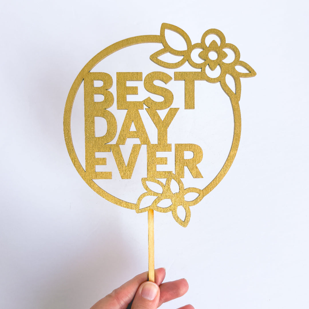 Gold 'Best Day Ever' cake topper held by a hand on a white background