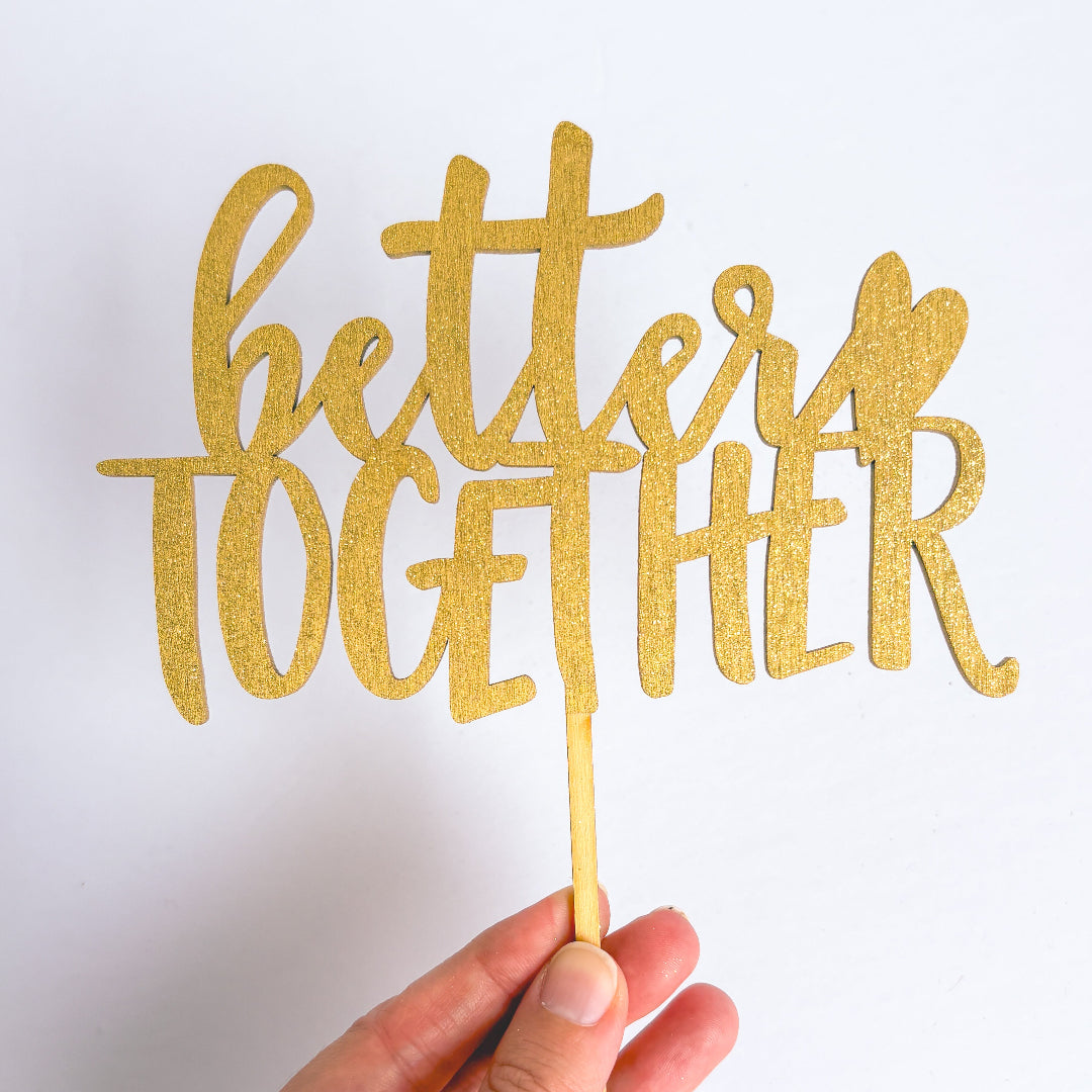 Gold glittery 'better together' sign held by a hand on a white background