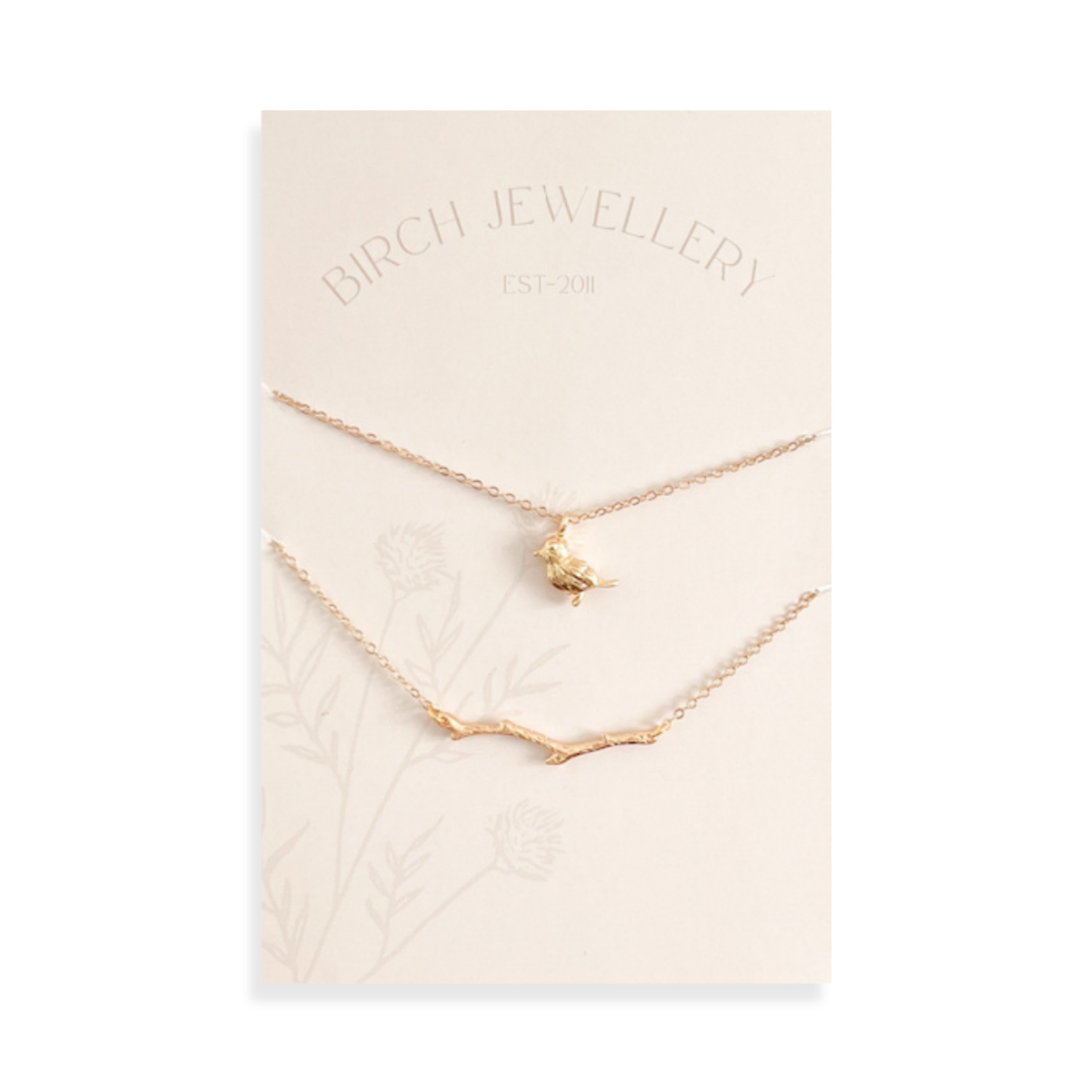 A pair of gold necklaces on a white card. The longer necklace features a branch pendant, and the shorter necklace features a bird pendant. 