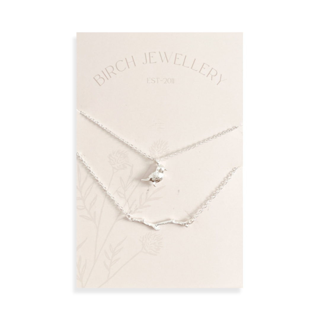 A pair of silver necklaces on a white card. The longer necklace features a branch pendant, and the shorter necklace features a bird pendant. 
