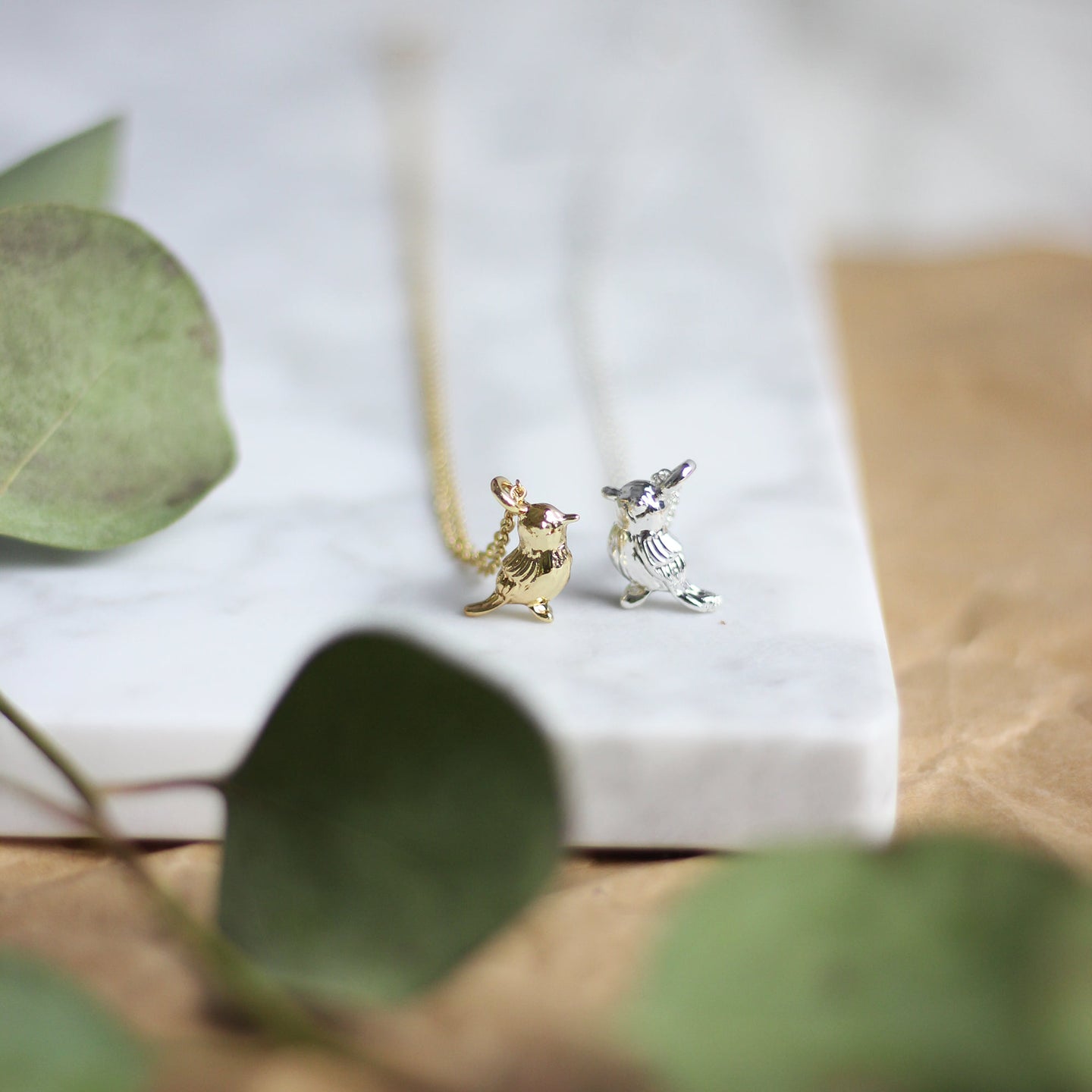 Two necklaces with bird pendants on a white marble surface. One necklace has a gold bird pendant, and the other has a silver bird pendant.