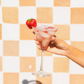 Hand holding a cocktail glass with ice and a strawberry against a checkered background