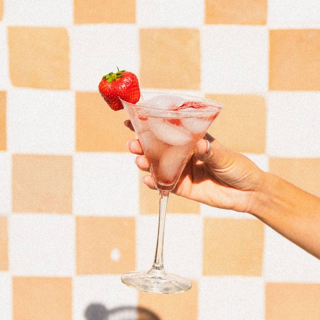 Hand holding a cocktail glass with ice and a strawberry against a checkered background