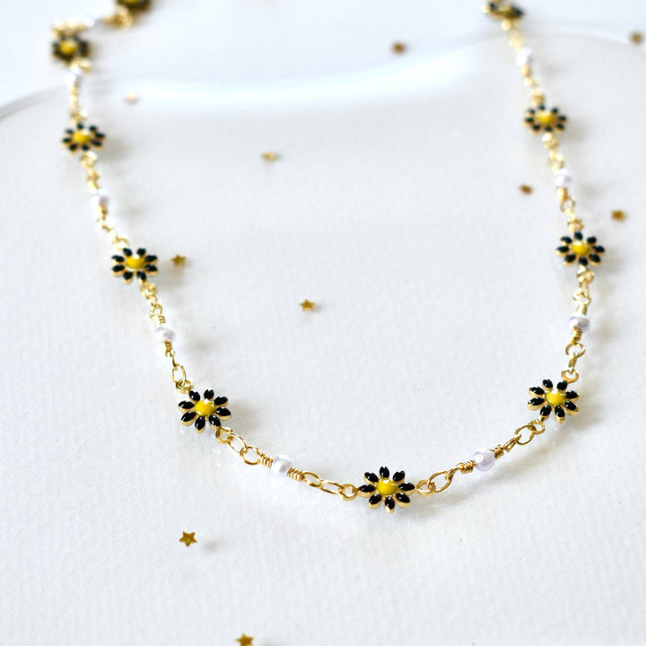A delicate gold necklace featuring a repeating pattern of small, yellow enamel daisies alternating with tiny white pearls. The necklace is lying on a white surface with gold glitter sprinkled around it.