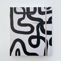A white-coloured agenda with a black abstract design on the cover. The word "AGENDA" is printed in gold foil on the cover, along with the brand name "IDLEWILD CO." The agenda has a white elastic band to keep it closed.
