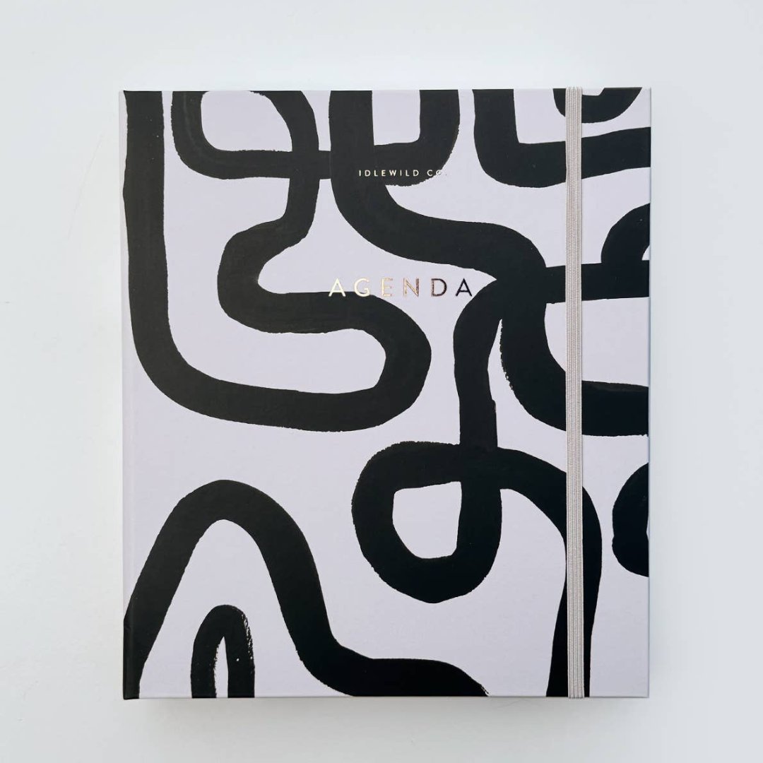 A white-coloured agenda with a black abstract design on the cover. The word "AGENDA" is printed in gold foil on the cover, along with the brand name "IDLEWILD CO." The agenda has a white elastic band to keep it closed.