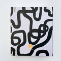 A white-coloured agenda with a black abstract design on the cover. The word "AGENDA" is printed in gold foil on the cover, along with the brand name "IDLEWILD CO." The agenda has a white elastic band to keep it closed.