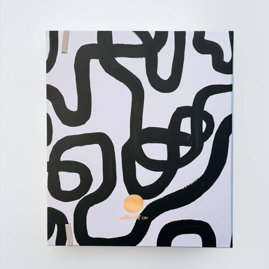 A white-coloured agenda with a black abstract design on the cover. The word "AGENDA" is printed in gold foil on the cover, along with the brand name "IDLEWILD CO." The agenda has a white elastic band to keep it closed.