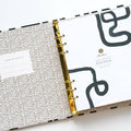 A white-coloured agenda with a black abstract design on the cover. The word "AGENDA" is printed in gold foil on the cover, along with the brand name "IDLEWILD CO." The agenda has a white elastic band to keep it closed.