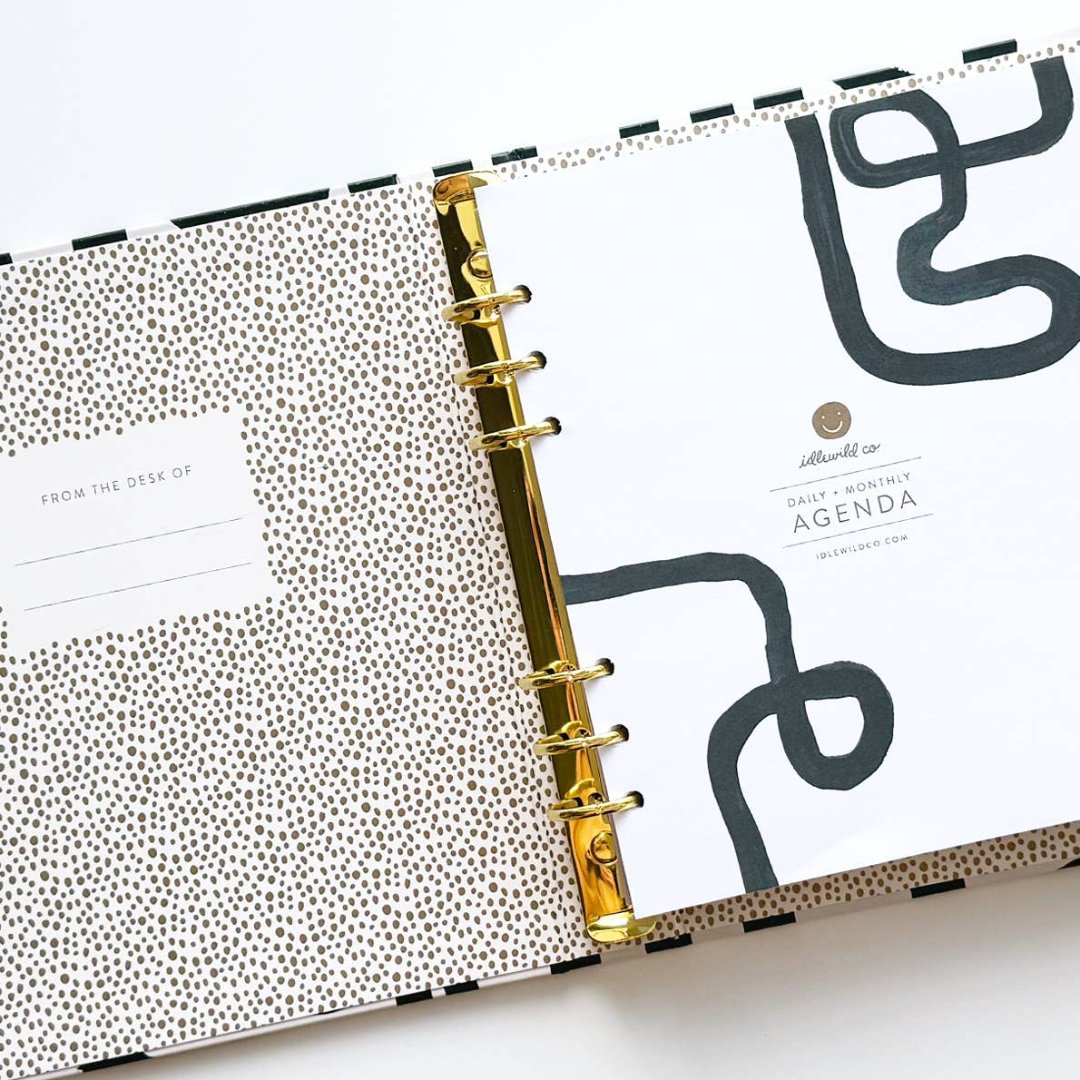 A white-coloured agenda with a black abstract design on the cover. The word "AGENDA" is printed in gold foil on the cover, along with the brand name "IDLEWILD CO." The agenda has a white elastic band to keep it closed.