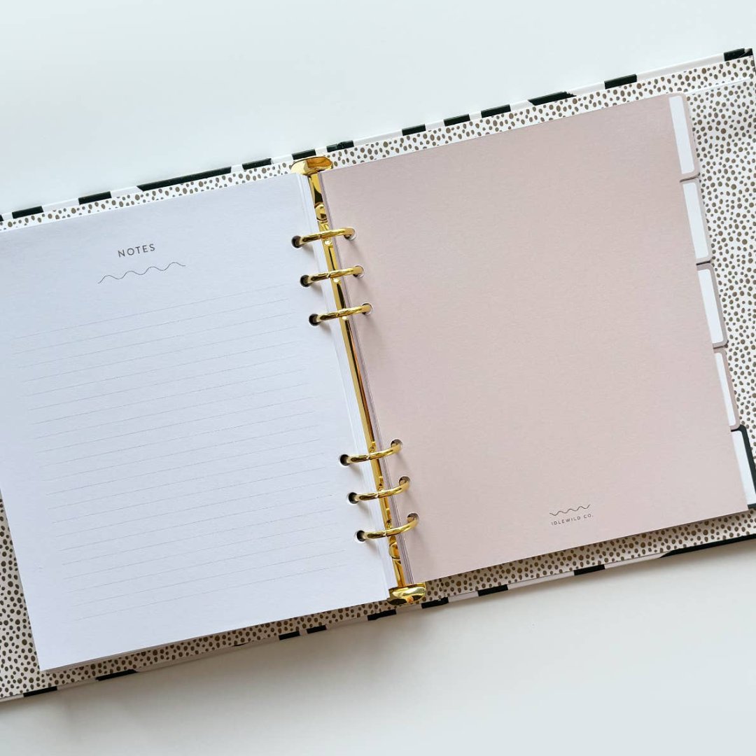 A white-coloured agenda with a black abstract design on the cover. The word "AGENDA" is printed in gold foil on the cover, along with the brand name "IDLEWILD CO." The agenda has a white elastic band to keep it closed.