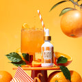 A bottle of Bliss Spritz citrus cooling & hydrating spray stands on a small orange pedestal beside a glass of orange drink with a striped straw, surrounded by oranges, citrus slices, and green leaves against a bright orange background.