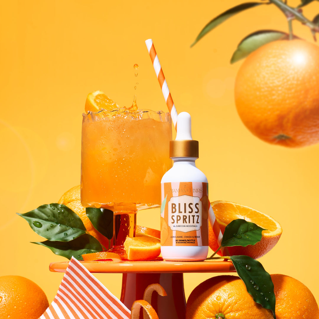 A bottle of Bliss Spritz citrus cooling & hydrating spray stands on a small orange pedestal beside a glass of orange drink with a striped straw, surrounded by oranges, citrus slices, and green leaves against a bright orange background.