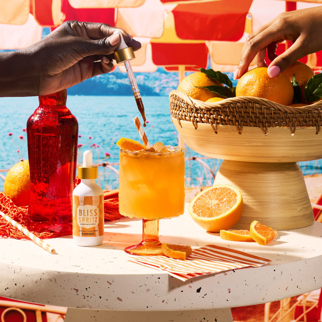 A styled outdoor scene with a hand holding a dropper above a glass of orange drink, surrounded by oranges, a red bottle, a small white bottle labeled Bliss Spritz, and a woven basket of oranges on a table with a beach background.