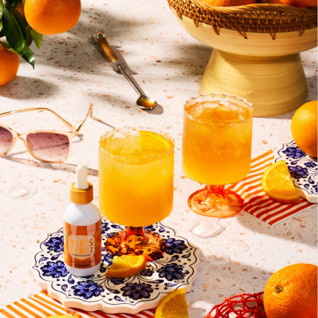 Two glasses of orange juice with a bottle of juice on a table with oranges and sunglasses.