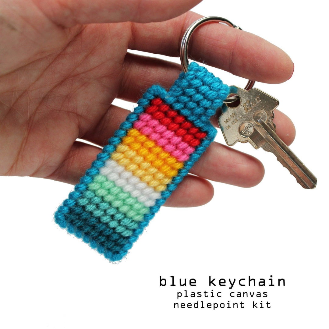 A hand holds a finished needlepoint keychain and a set of keys. The keychain is rectangular with a striped pattern in shades of blue, green, yellow, pink, and red, and is attached to a silver key ring.
