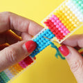 A hand holds a finished needlepoint keychain and a set of keys. The keychain is rectangular with a striped pattern in shades of blue, green, yellow, pink, and red, and is attached to a silver key ring.