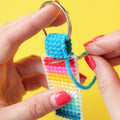 A hand holds a finished needlepoint keychain and a set of keys. The keychain is rectangular with a striped pattern in shades of blue, green, yellow, pink, and red, and is attached to a silver key ring.