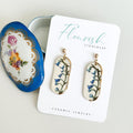 A pair of small, elongated oval dangle earrings with a white base and delicate bluebell flower imprints in shades of blue and green, accented with a touch of luxurious 22K gold detailing around the edges. They hang from simple 14K gold plated ball posts