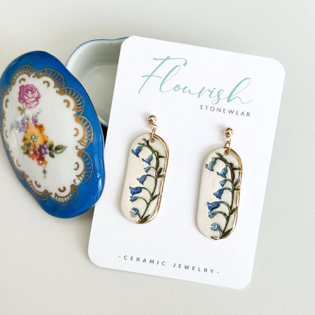 A pair of small, elongated oval dangle earrings with a white base and delicate bluebell flower imprints in shades of blue and green, accented with a touch of luxurious 22K gold detailing around the edges. They hang from simple 14K gold plated ball posts