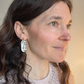 A pair of small, elongated oval dangle earrings with a white base and delicate bluebell flower imprints in shades of blue and green, accented with a touch of luxurious 22K gold detailing around the edges. They hang from simple 14K gold plated ball posts