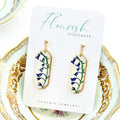 A pair of small, elongated oval dangle earrings with a white base and delicate bluebell flower imprints in shades of blue and green, accented with a touch of luxurious 22K gold detailing around the edges. They hang from simple 14K gold plated ball posts