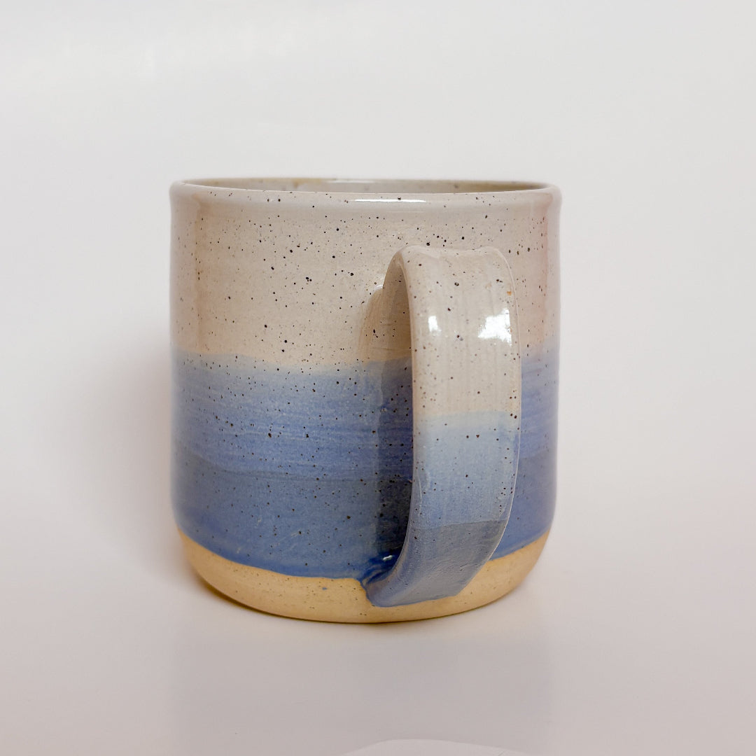 A handmade ceramic mug with a blue ombre glaze, featuring a speckled texture, displayed on a light gray background. The mug has a large, rounded handle and is made in Mississippi Mills, Ontario, Canada.