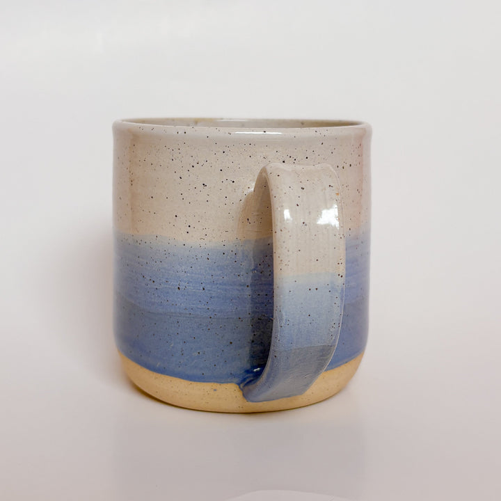 A handmade ceramic mug with a blue ombre glaze, featuring a speckled texture, displayed on a light gray background. The mug has a large, rounded handle and is made in Mississippi Mills, Ontario, Canada.