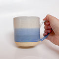 A handmade ceramic mug with a blue ombre glaze, featuring a speckled texture, displayed on a light gray background. The mug has a large, rounded handle and is made in Mississippi Mills, Ontario, Canada.