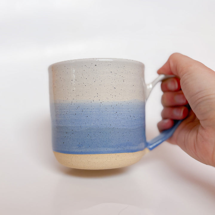 A handmade ceramic mug with a blue ombre glaze, featuring a speckled texture, displayed on a light gray background. The mug has a large, rounded handle and is made in Mississippi Mills, Ontario, Canada.