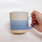 A handmade ceramic mug with a blue ombre glaze, featuring a speckled texture, displayed on a light gray background. The mug has a large, rounded handle and is made in Mississippi Mills, Ontario, Canada.
