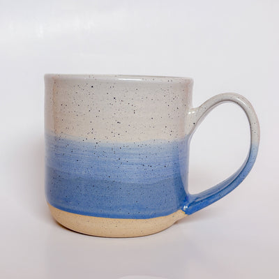 A handmade ceramic mug with a blue ombre glaze, featuring a speckled texture, displayed on a light gray background. The mug has a large, rounded handle and is made in Mississippi Mills, Ontario, Canada.